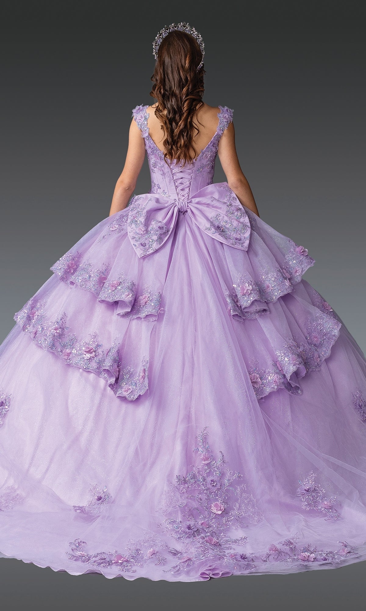 A woman with long brown hair, shown from the back, wears the Quinceanera Dress 1952 By Dancing Queen—a lilac gown with floral lace embroidery, layered skirts, a large back bow, and a jeweled headpiece.